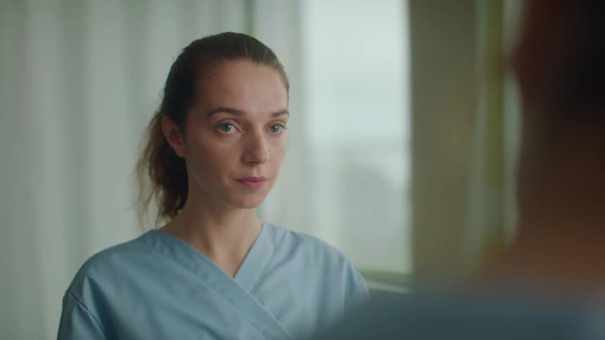 Young woman wearing blue scrubs listening to a doctor inside a hospital room