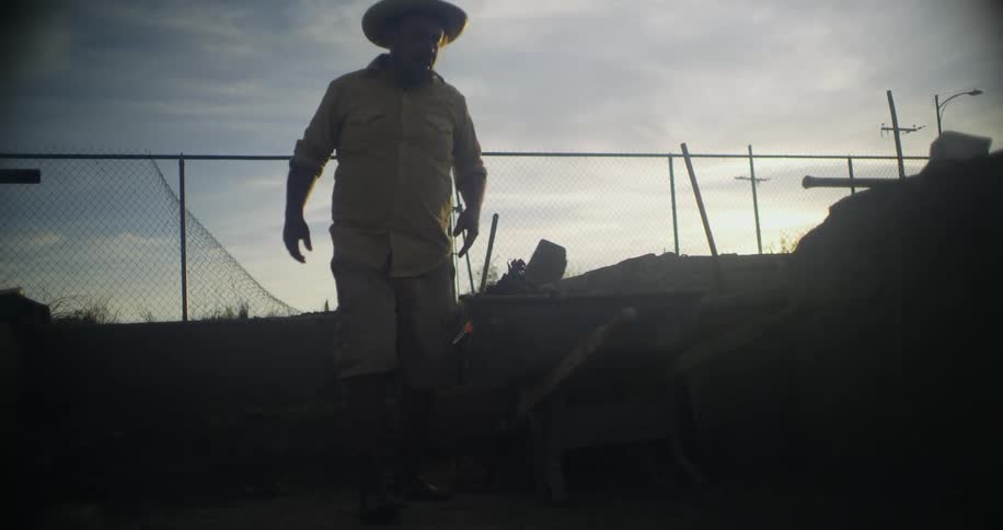 Man works in shadow to dig up dirt to place in wheel barrow under cloudy sky