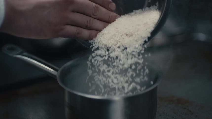 A chef is pouring rice into a pot of boiling water in a commercial kitchen during the daytime