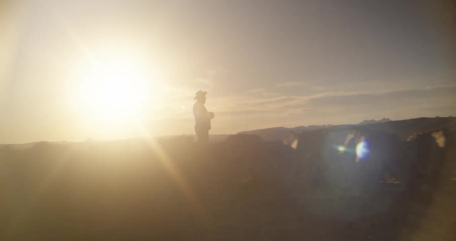Man with cowboy hat is silhouetted against the sunset standing still in field