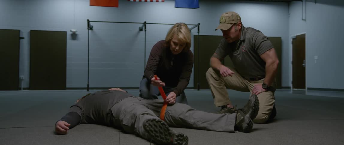 A woman ties a belt around a man's leg who is laying down