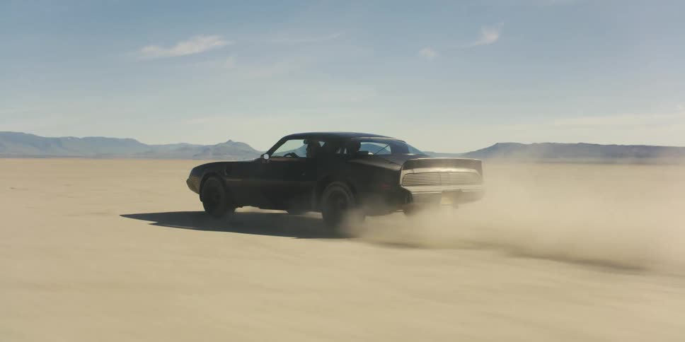A black trans am travels across a desert with mountains in the distance