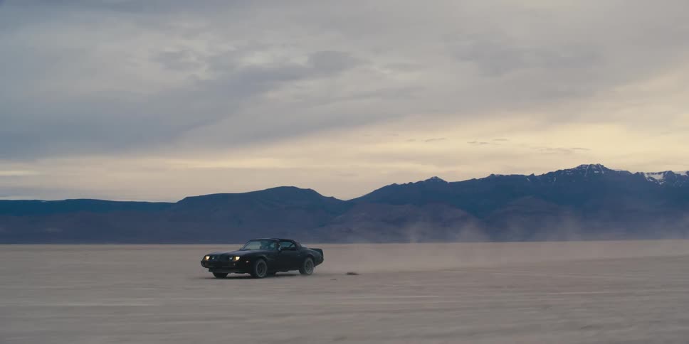 A car driving on a desert with a mountain range the sky and clouds in the background