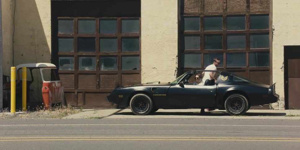 A black trans am with ttops is traveling near buildings when it stops and two people get out and change drivers