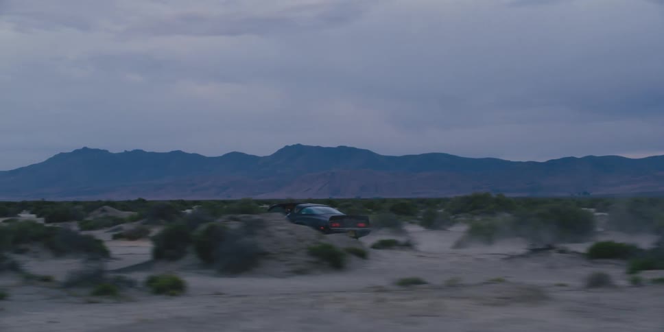Car drives across sand between clumps of vegetation kicking up dust as it passes dunes with mountains in background under cloudy sky