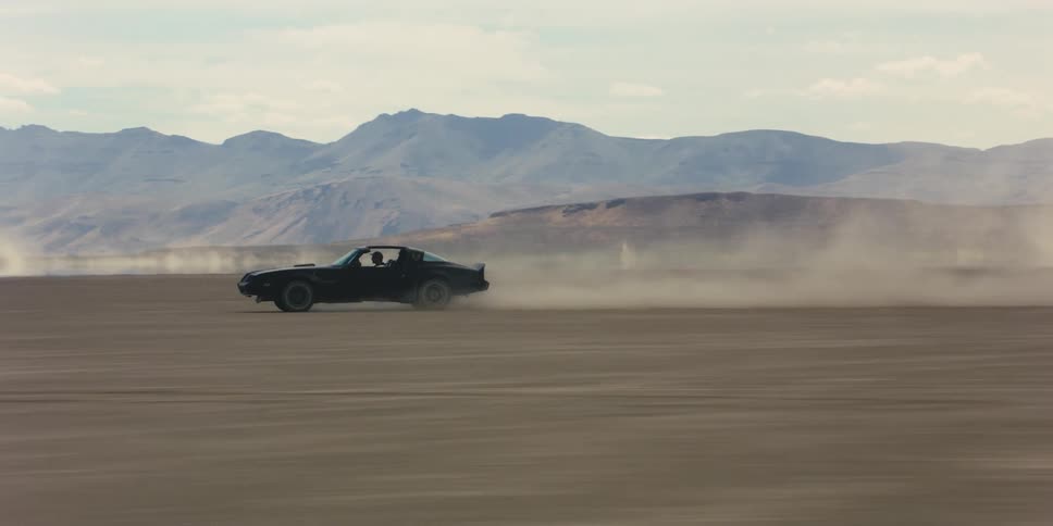 A car driving on a desert with a mountain range the sky and clouds in the background