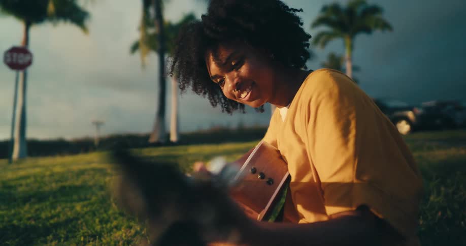 A woman is sitting in the grass of a public park and playing a guitar in the evening with storm clouds in the background