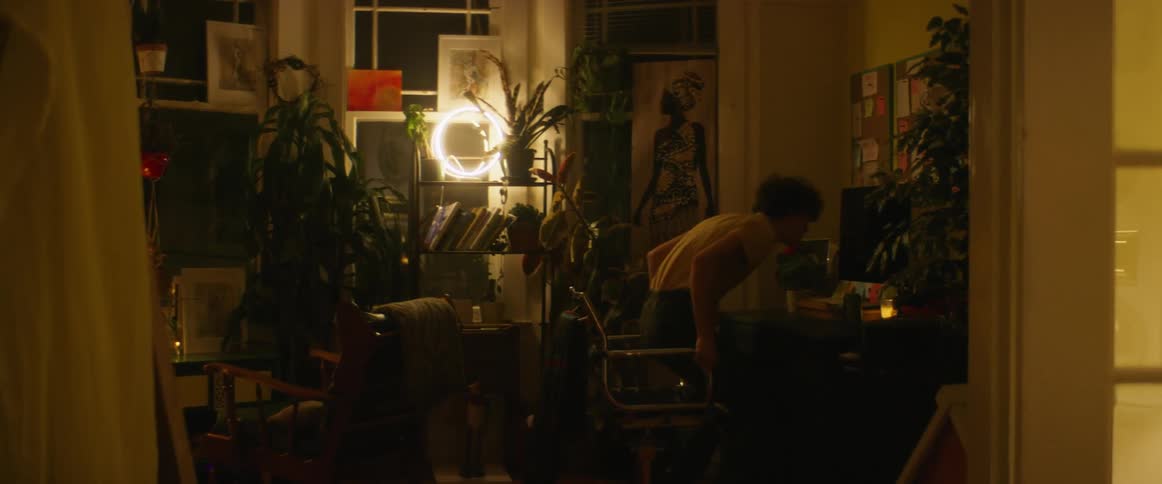 Man with wavy hair shirt and black pants drinks beverage and sits down in chair to turn on computer in dim lit room