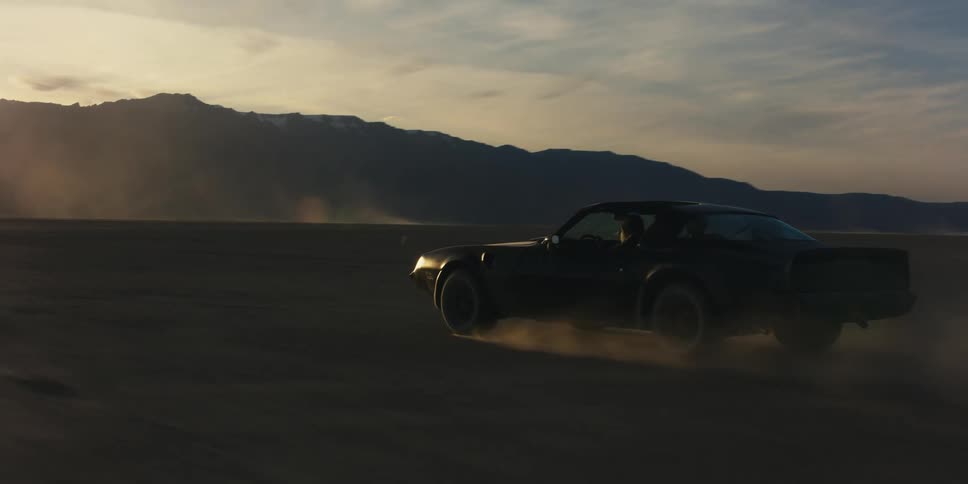 Woman driver and male passenger speed across desert kicking up dust as they pass through dust filled air with mountains in background