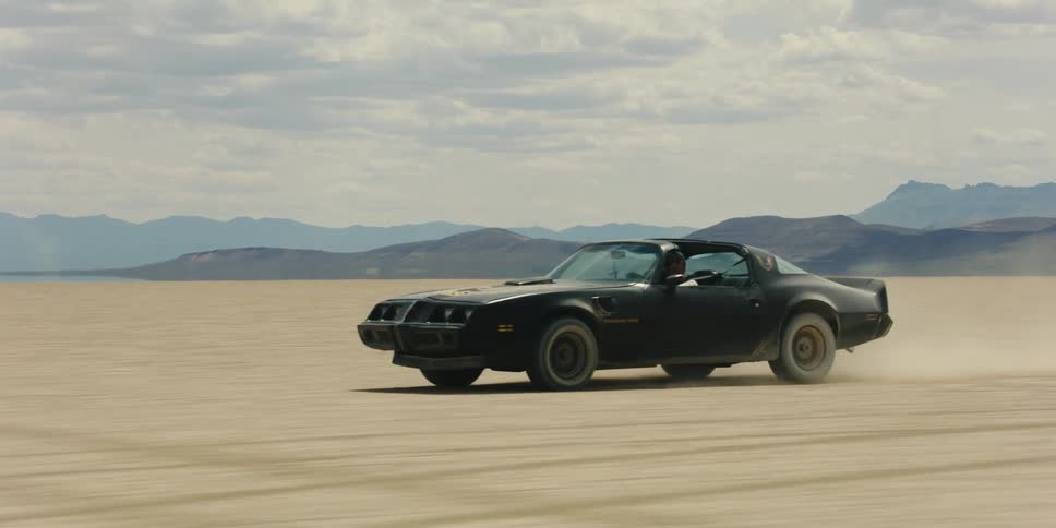 Person drives sports car across desert kicking up dust with mountains on horizon under cloudy sky