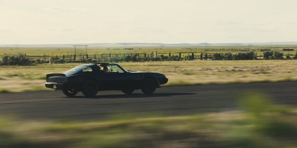 Two people drive along highway through countryside past pastures in sports car