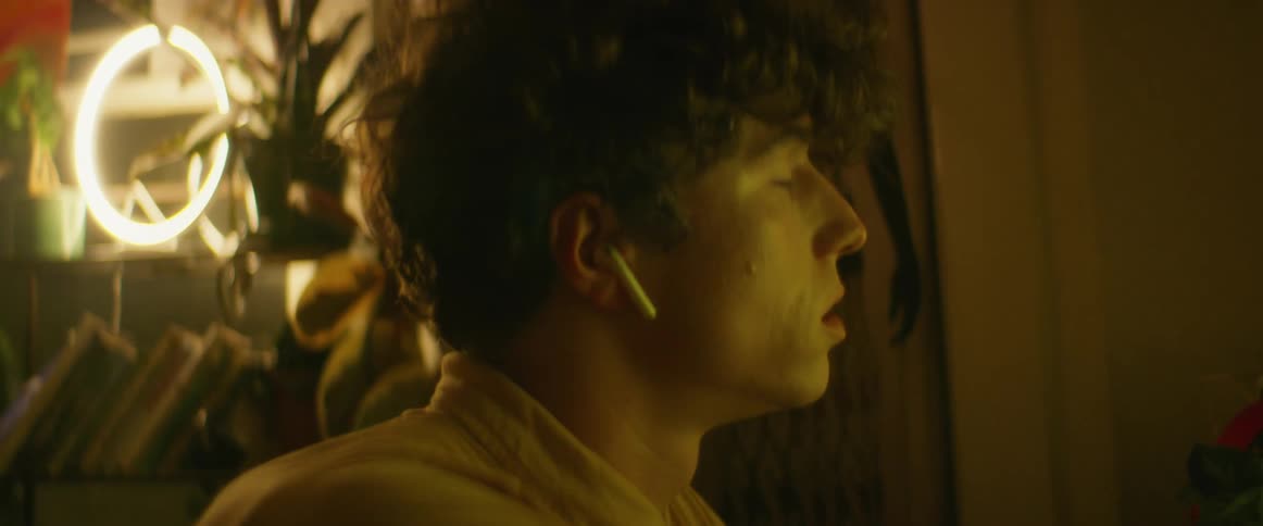 Man with curly hair yellow shirt sits at desk wearing earphones and using computer