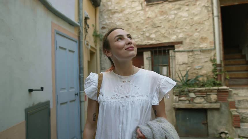 A woman wearing a white dress and carrying a jacket is walking in an alley looking at buildings and smiling