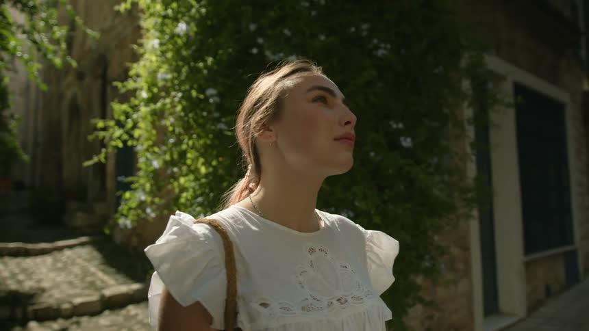A woman stands in a narrow street and looks up at old stone buildings