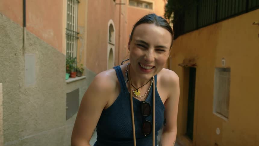A woman laughs while posing in an alley between buildings while holding a camera
