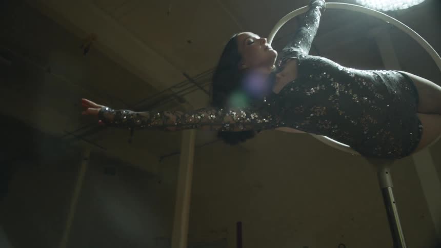 A woman in a dance studio is dressed in a sparkling leotard while hanging from a ring pole at night