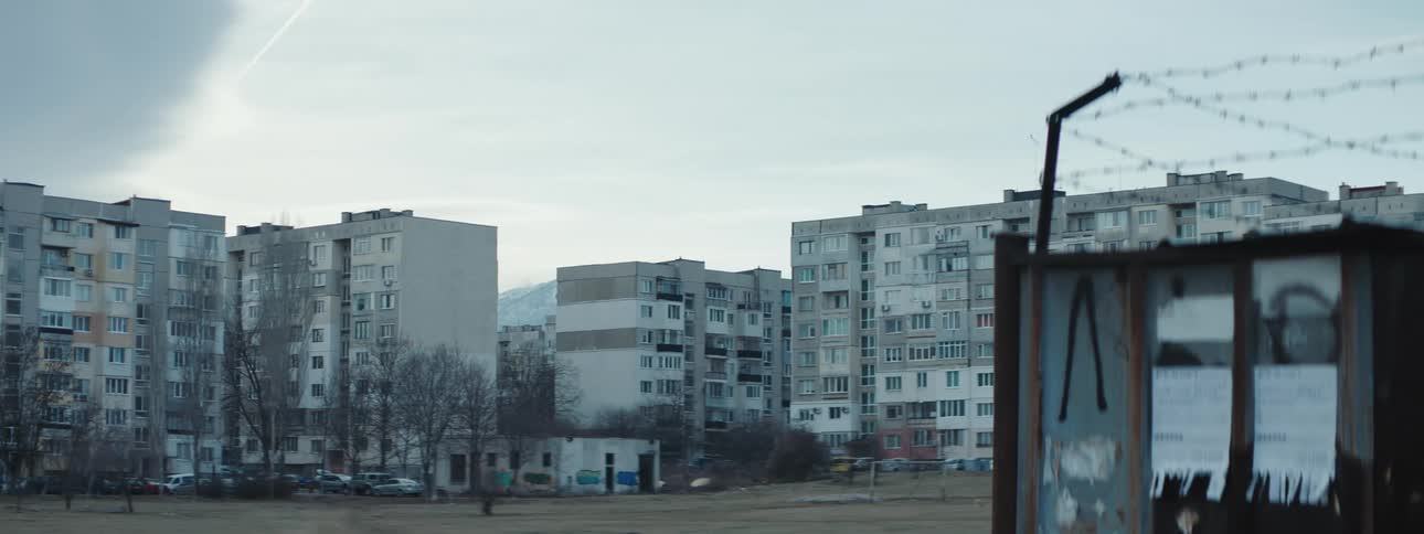 Traffic moves along road past apartment buildings across field from barbed wire enclosed area