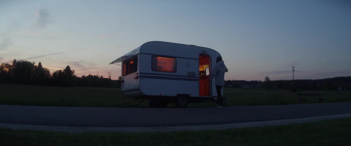 A person wearing a light colored jacket walks across a road to a trailer with an open door and a light on inside