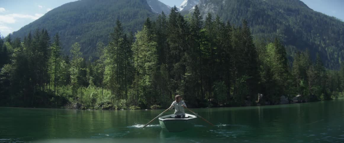 A man is rowing a boat across a lake toward a forest with mountains in the background during the day