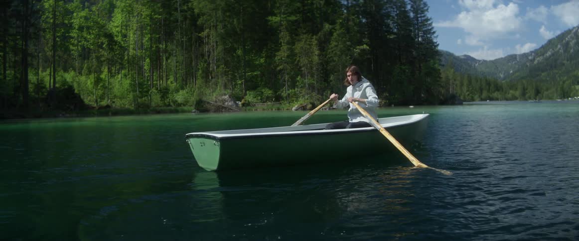 Man rows boat across rippling water with forest and mountains in background