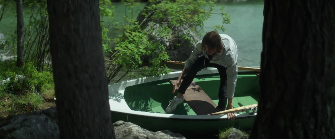 A young man in a rowboat lands on the shore and climbs out of the boat