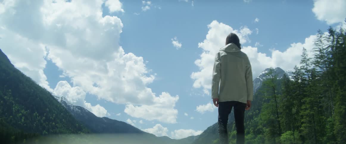 A man walking by a lake with a mountain range the sky and clouds in the background