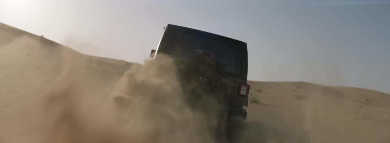 Vehicle spins tires kicking up sand as it drives up desert slope under sunny sky