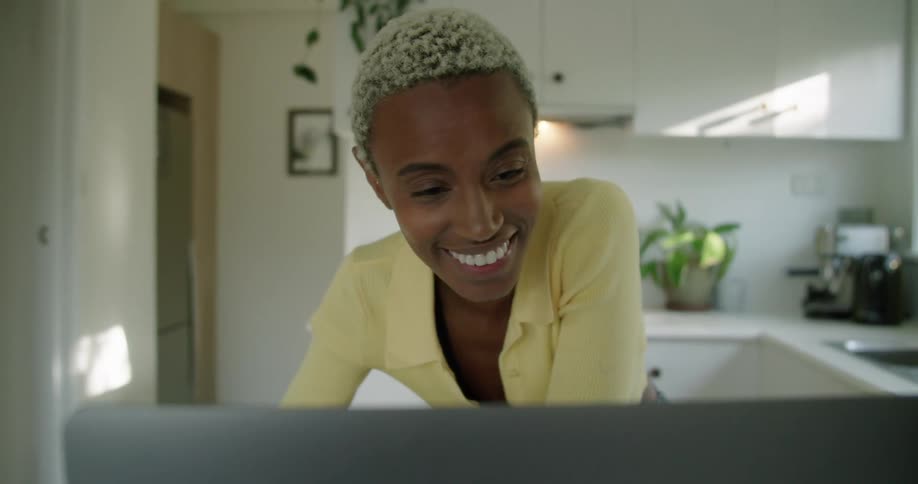 A woman is standing in her kitchen smiling at a laptop