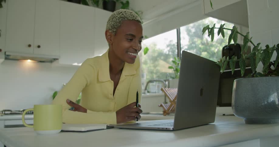 A woman is in a kitchen talking and waving at a laptop