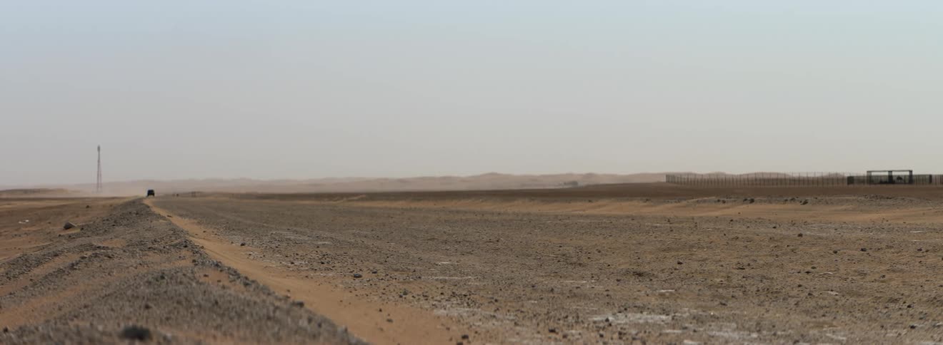 Vehicle drives along dirt road kicking up dust approaching fenced pasture with mountains on horizon