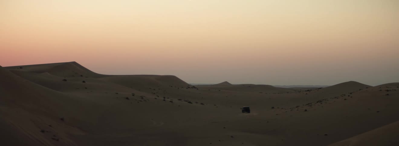 A vehicle driving in a desert with dunes with the sky and clouds in the background