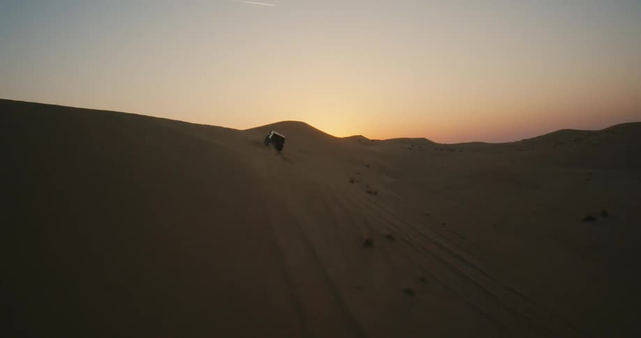 An aerial view of a vehicle driving over sand dunes in a desert at sunset