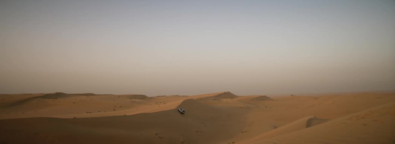 A vehicle driving in a desert landscape with dunes with the sky and clouds in the background