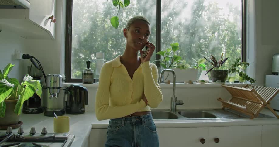 A woman standing in a kitchen talking on a smart phone with a sink and a window in the background