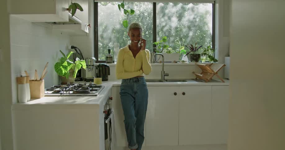 A woman is standing in her home kitchen making a phone call on her smartphone on a breezy day