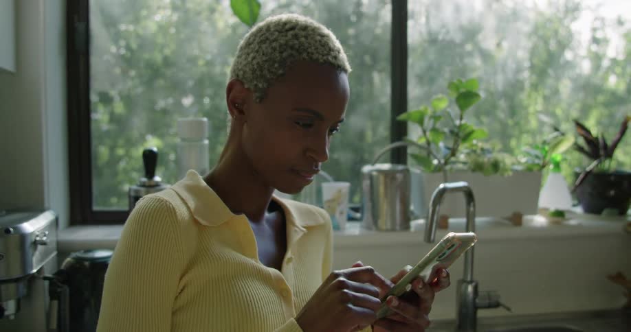 Woman with short hair and yellow vest smiles and scrolls up and down on her smartphone screen in the kitchen