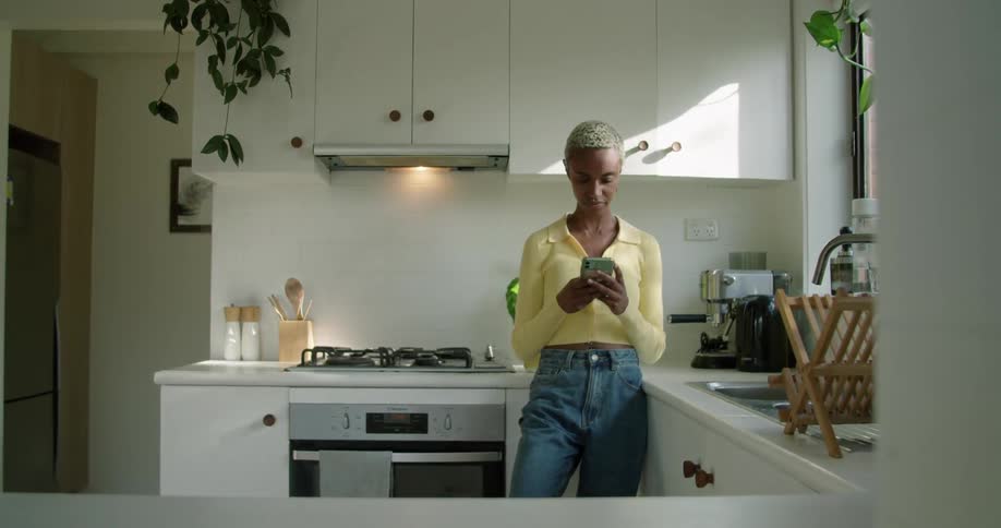 Woman leans against kitchen counter near stove scrolling on mobile phone