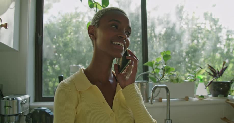 A woman standing in a kitchen talking on a smart phone with a sink and a window in the background