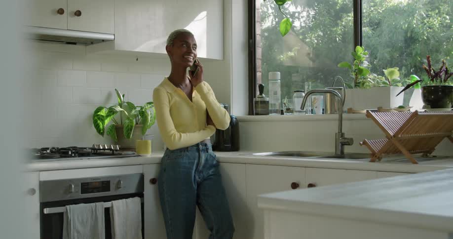 A woman is standing in a kitchen smiling as she talks on her cell phone