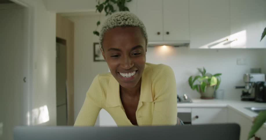 A woman is smiling and waving while video chatting on a laptop in her kitchen in the daytime