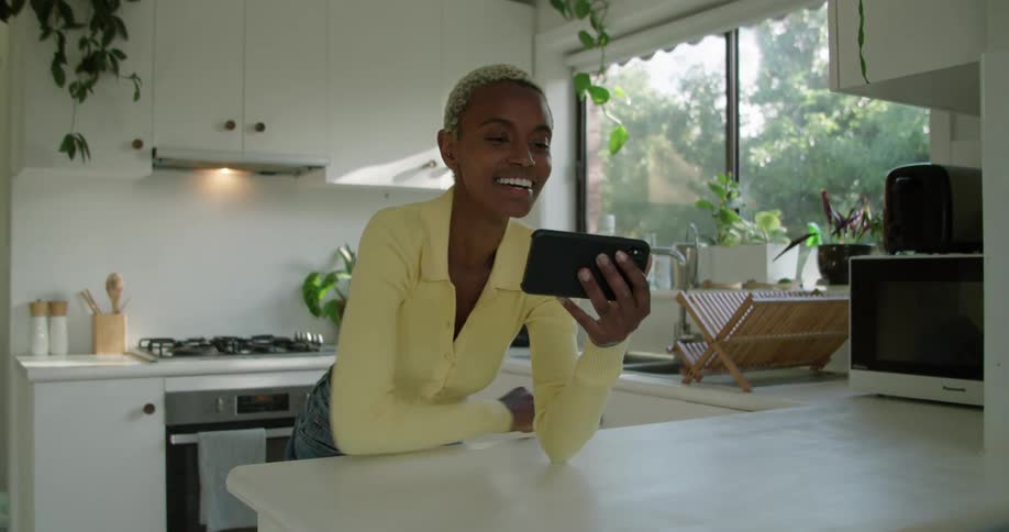 A woman stands in her kitchen and waves and laughs as she video chats on her cell phone
