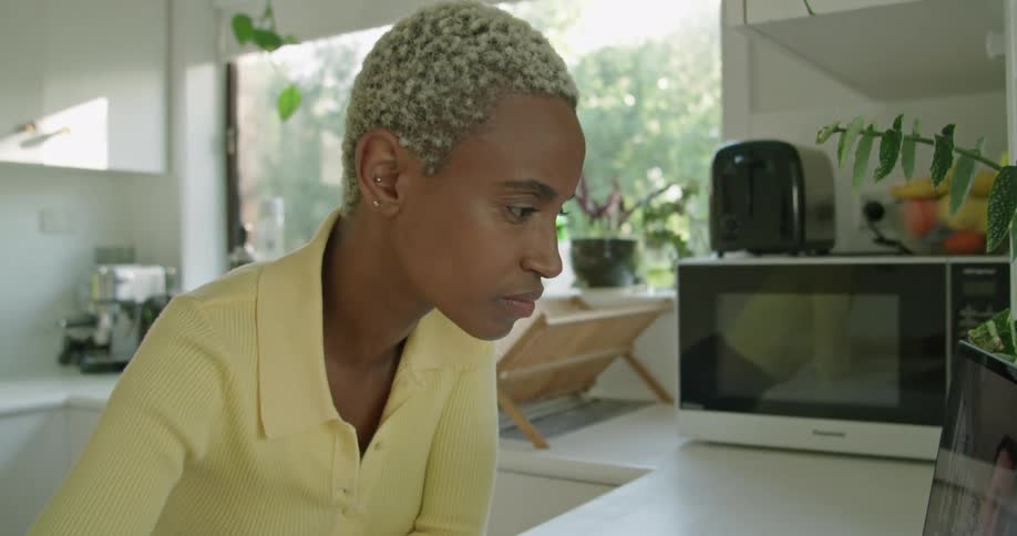 Woman with short hair and yellow shirt makes video call on laptop and writes in notebook leaning on kitchen island