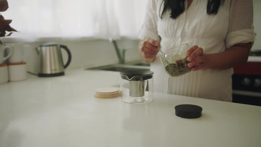 A woman wearing a white blouse is in a kitchen making coffee on a kitchen counter