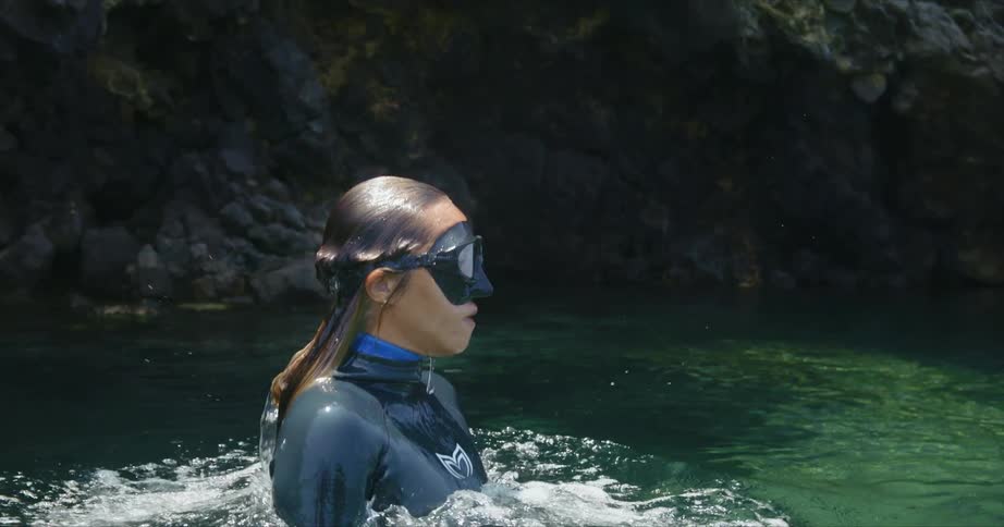 A woman wearing swimming goggles and a wet suit emerges from underwater