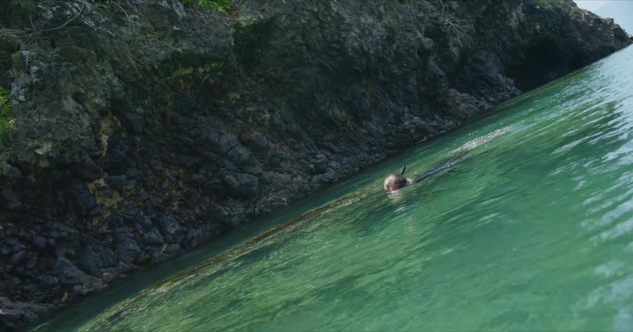 Woman with brown hair mask snorkel wetsuit and flippers swims in water next to cliff