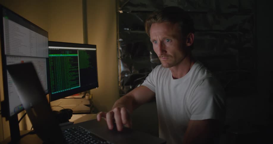 A man is sitting in front of computer monitor screens and typing on a laptop