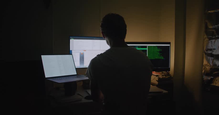 A man in a room by a desk with laptops and computers with a wall in the background