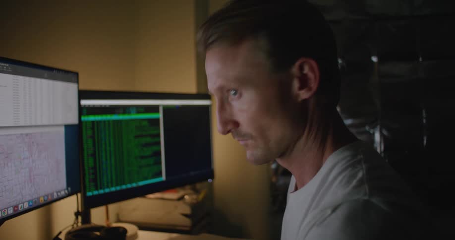 A man in a room sitting in front of a desk with a laptop and computers with a wall in the background