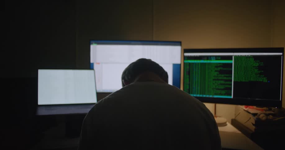 A man is sitting in front of a laptop computer and two other computer monitors