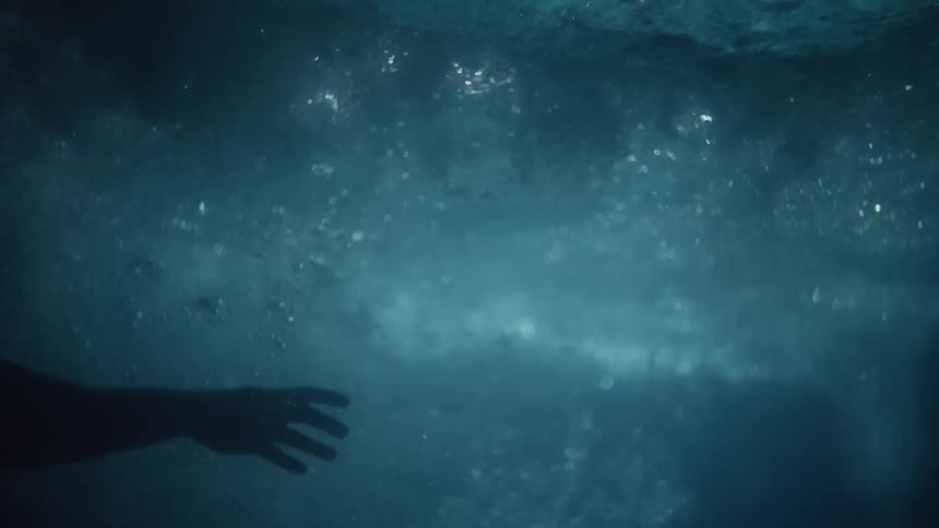 Persons silhouette floats underwater as wave crashes creating bubbles and letting light shine through churning water